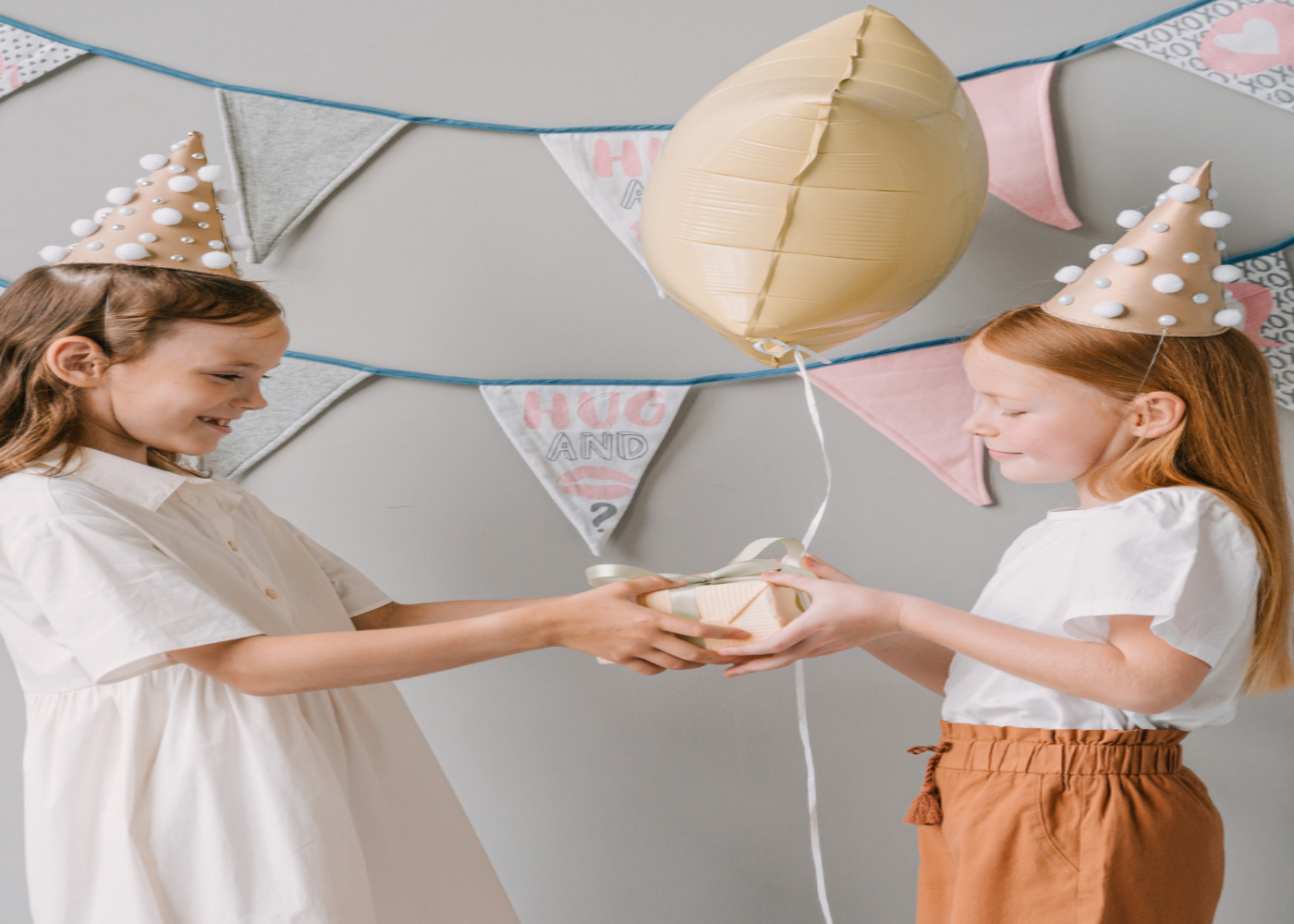 Two children wearing party hats holding a beige balloon with decorative flags in the background. Representing Gift Collection at The Vintage Teddy Children's Boutique.