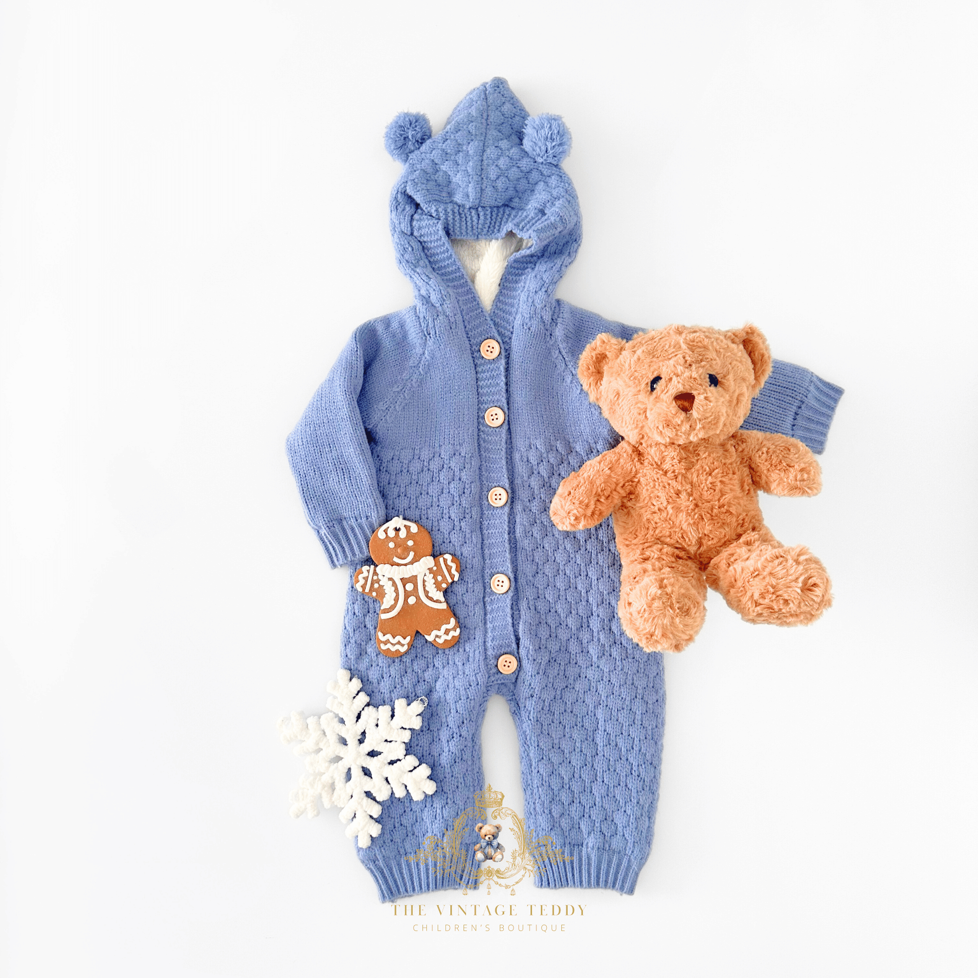 Baby boys knit hooded jumpsuit in cornflower blue with a textured cable-knit pattern, button closure, styled with a gingerbread man, and a plush white snowflake, and a cute brown teddy bear on a white background surface.