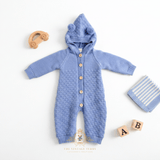 Cornflower blue knitted baby hoodie romper with button closure, laid flat on a white background styled with props including a wooden toy car, striped blanket, and A & B letter wood blocks. The brand name 'The Vintage Teddy' is visible at the bottom.