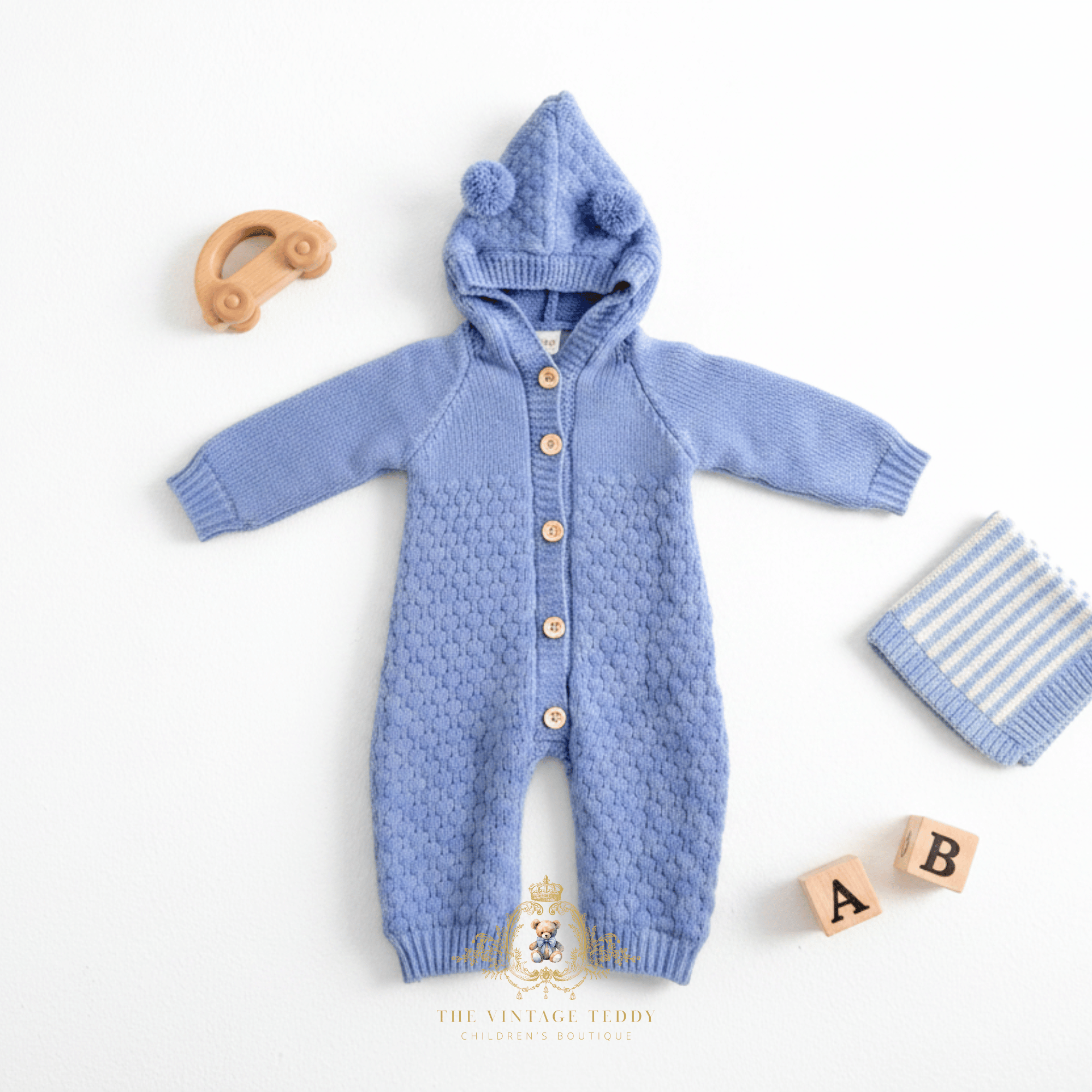 Cornflower blue knitted baby hoodie romper with button closure, laid flat on a white background styled with props including a wooden toy car, striped blanket, and A & B letter wood blocks. The brand name 'The Vintage Teddy' is visible at the bottom.