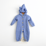 Cornflower blue knitted baby jumpsuit with a hood featuring two teddy bear pom-poms, front button closure, and a textured delicate pattern, displayed against a plain white background.