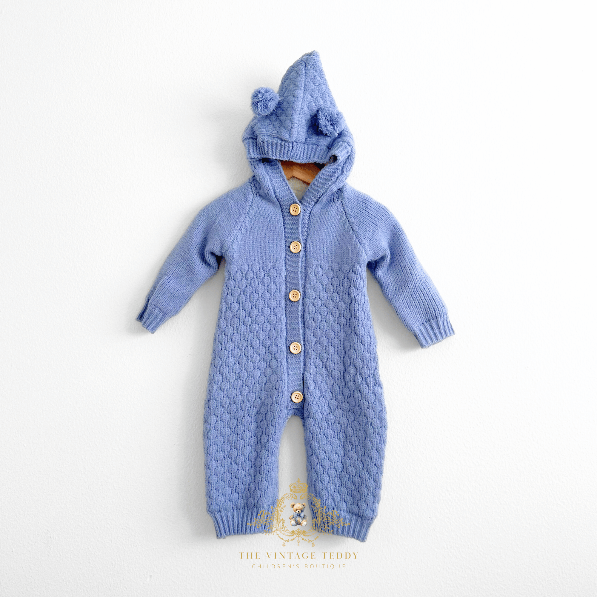 Cornflower blue knitted baby jumpsuit with a hood featuring two teddy bear pom-poms, front button closure, and a textured delicate pattern, displayed against a plain white background.