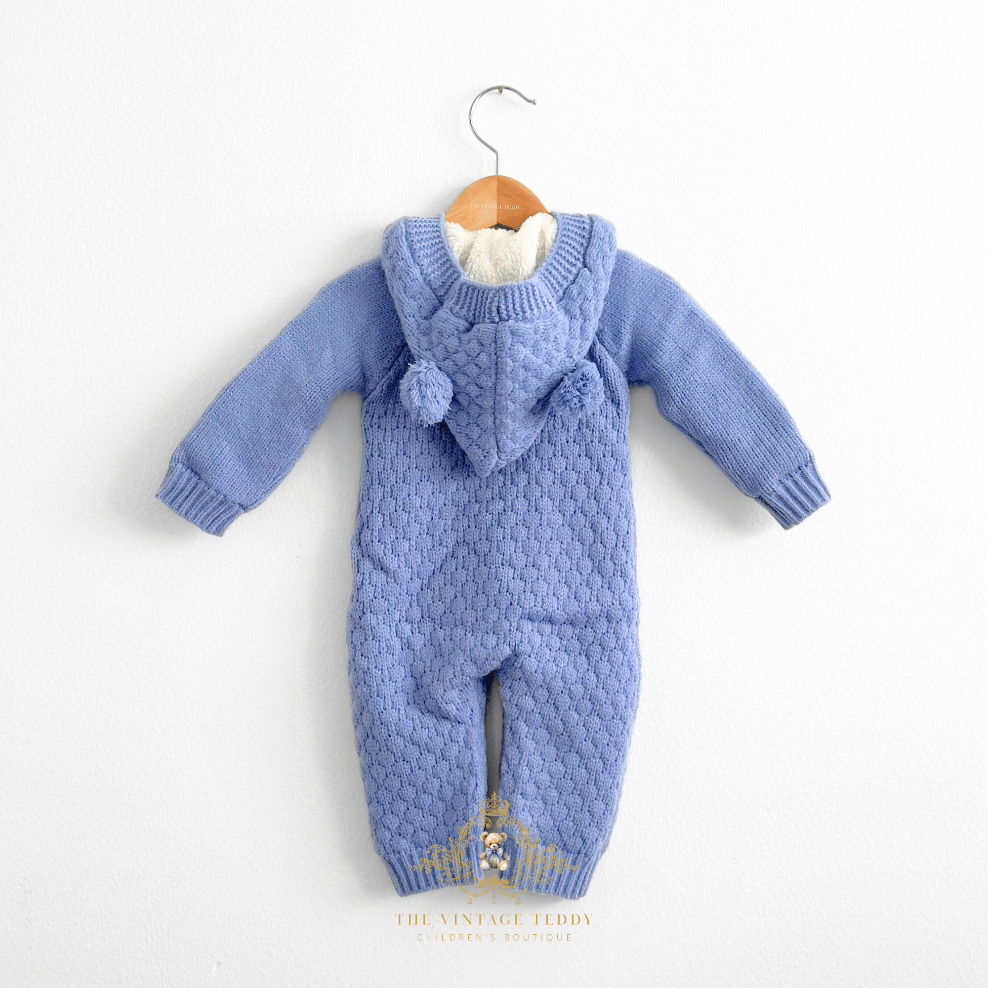 Cornflower blue knit baby one-piece  sweatsuit with a hood, textured cable-knit pattern, and faux-fur fleece lining, hanging on a wooden hanger against a white wall.