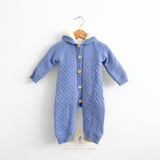 Baby boy hooded knit jumpsuit in cornflower blue with a white warm fleece sherpa lining and buttons down the center, hanging on a natural wood hanger against a white wall.