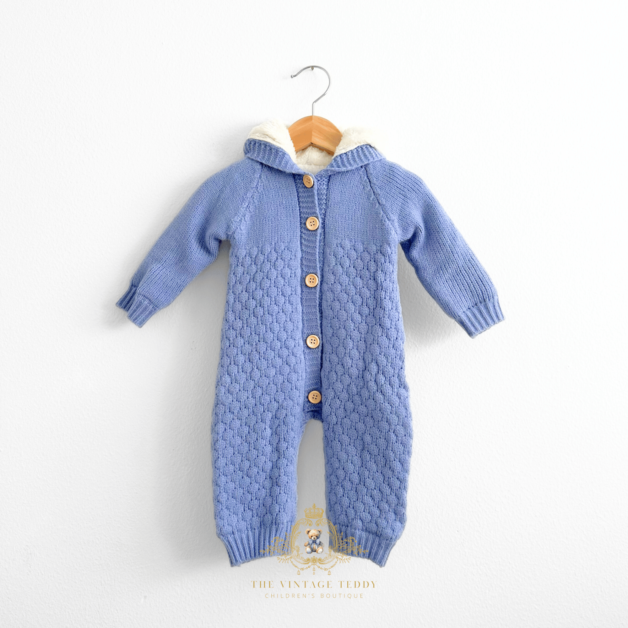 Baby boy hooded knit jumpsuit in cornflower blue with a white warm fleece sherpa lining and buttons down the center, hanging on a natural wood hanger against a white wall.