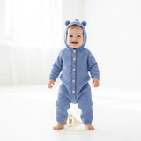 Baby boy wearing a cornflower blue knitted hooded one-piece outfit with buttons down the front, standing barefoot on a bright white studio floor. The image has a watermark at the bottom reading 'THE VINTAGE TEDDY CHILDREN'S BOUTIQUE'.