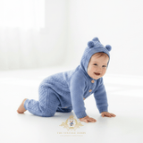 Baby boy wearing a cornflower blue knitted hoodie romper with bear ears on the hood, crawling on a bright white floor. The brand logo 'The Vintage Teddy' is visible at the bottom.