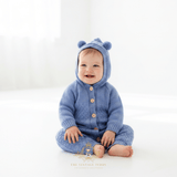 Baby boy sitting bare foot and smiling on a white floor wearing a blue knitted hooded romper with bear ears on the hood and buttons on the front, in a bright white studio setting.
