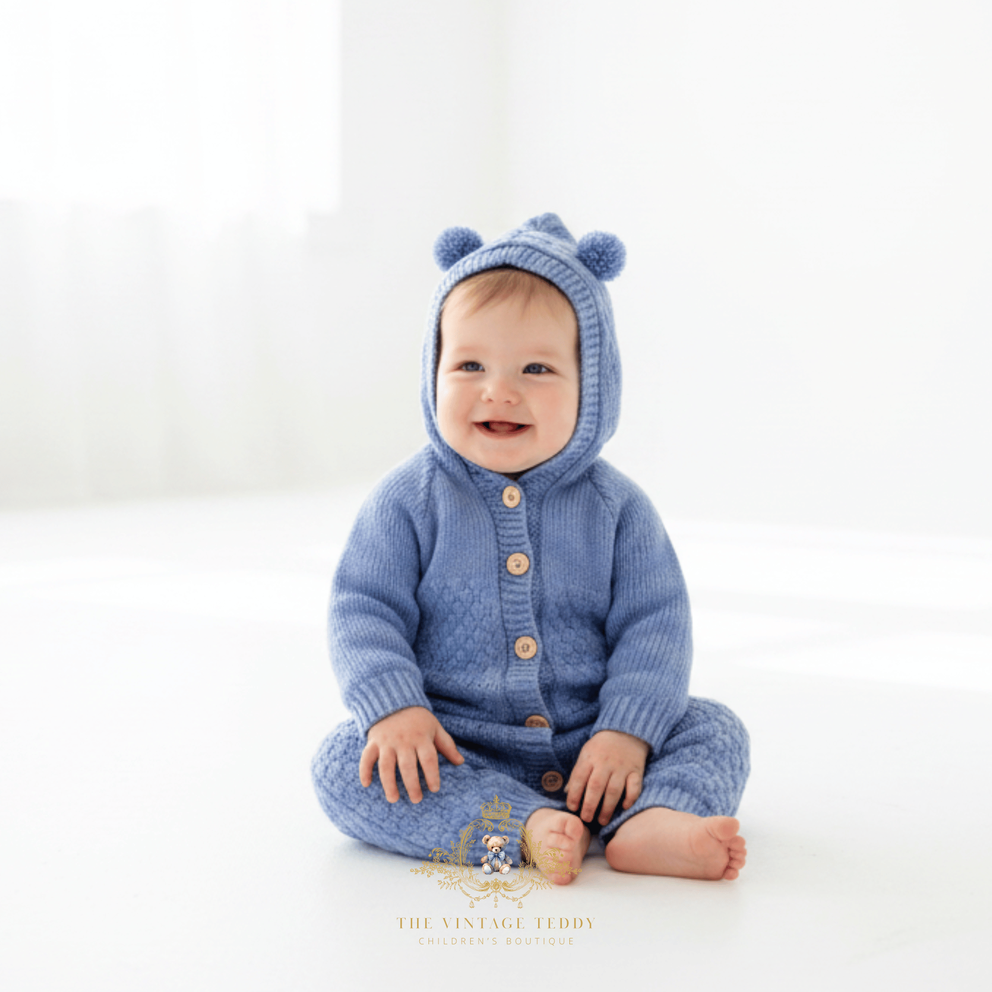 Baby boy sitting bare foot and smiling on a white floor wearing a blue knitted hooded romper with bear ears on the hood and buttons on the front, in a bright white studio setting.