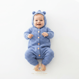 A baby boy wearing a light blue knitted hooded jumpsuit with bear ears on the hood, lying on a white background surface. The baby jumpsuit has a front button closure with tan buttons. The baby is bare foot and smiling.