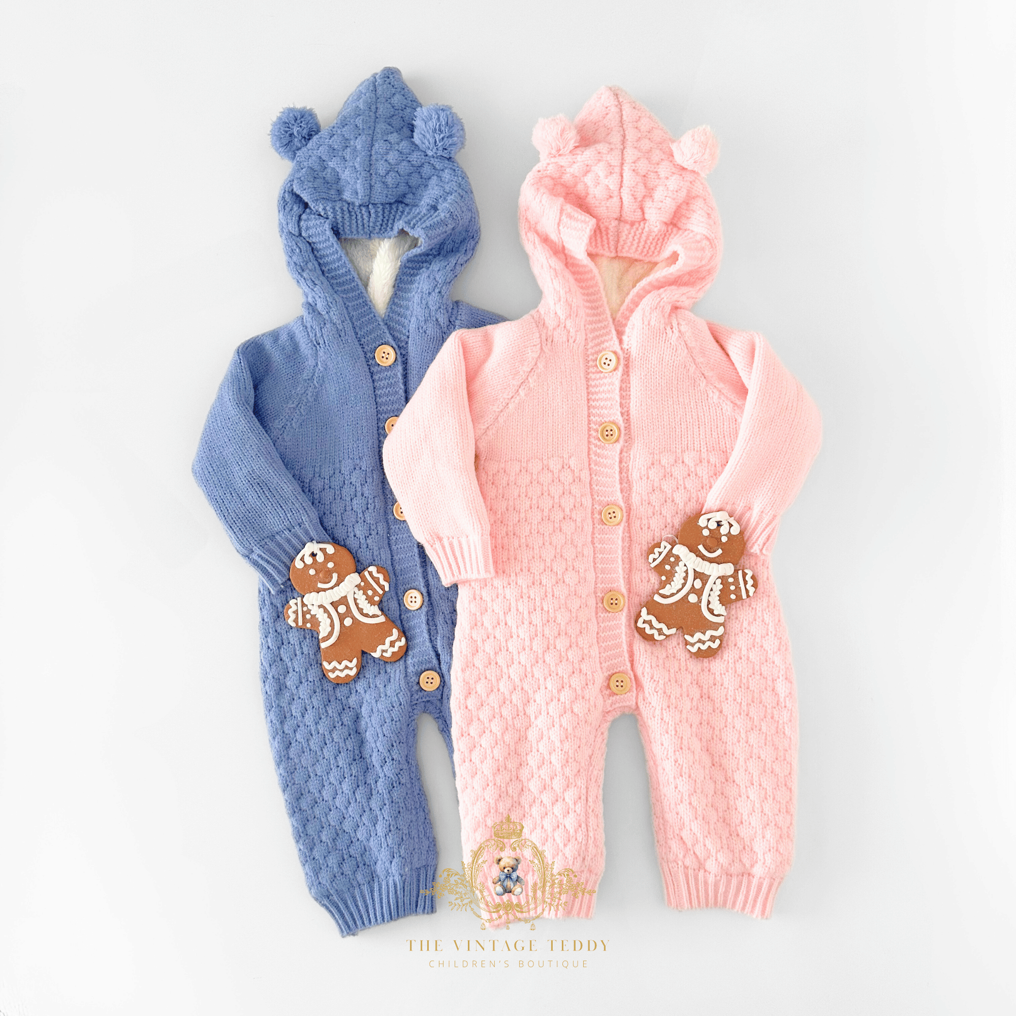 Two baby knit hooded one-piece outfits laid flat on a white surface background: one cornflower blue and one pastel pink, both with textured cable-knit patterns, button closures, bear-ear hoods, and styled with two gingerbread men ornaments laid on the lower front of each baby romper.