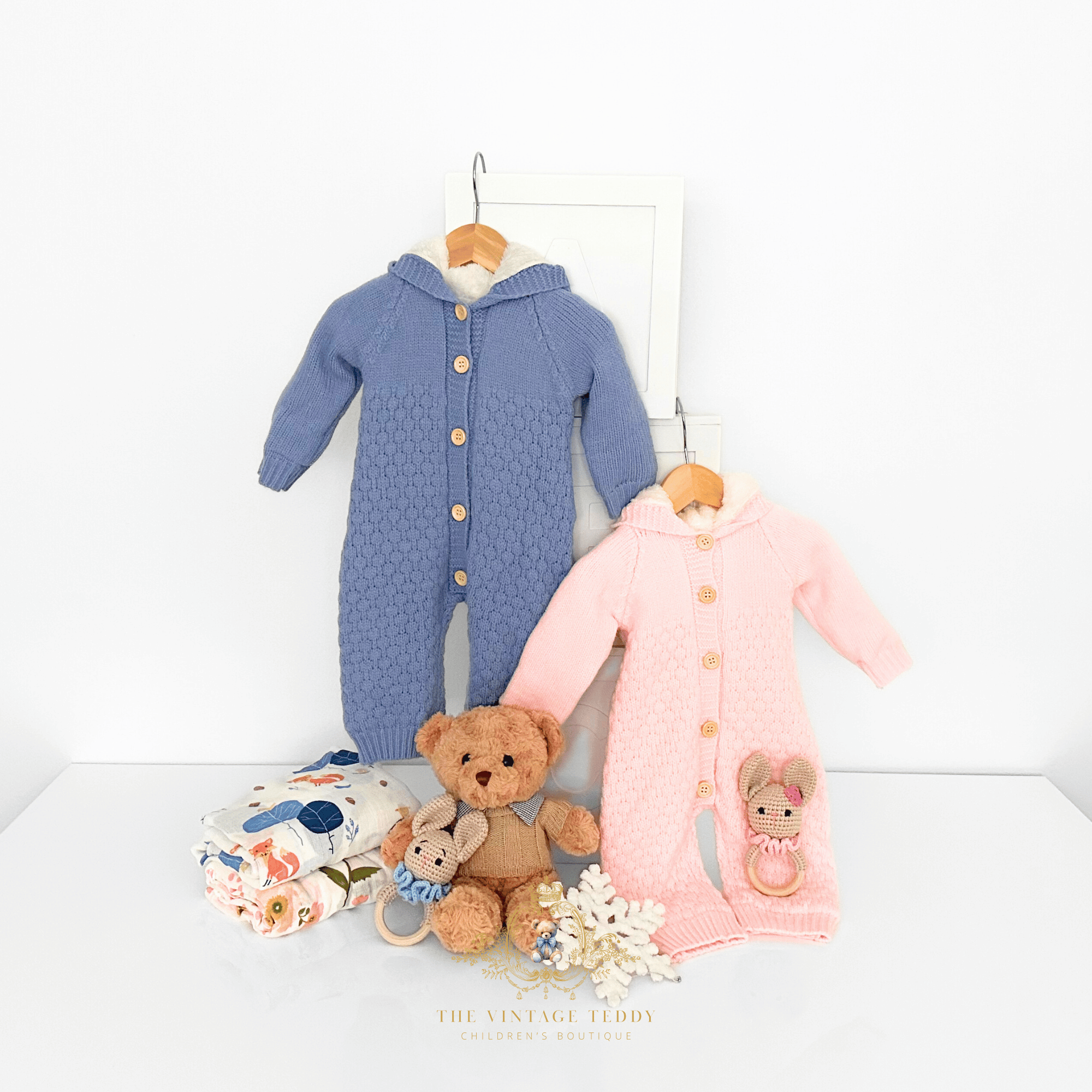 Two knitted baby one-piece rompers hanging on wooden hangers against a white wall on large ABC white wooden blocks, with a folded floral and woodland characters blankets, snowflake, teddy bears, and crochet bunny rattles arranged below. 