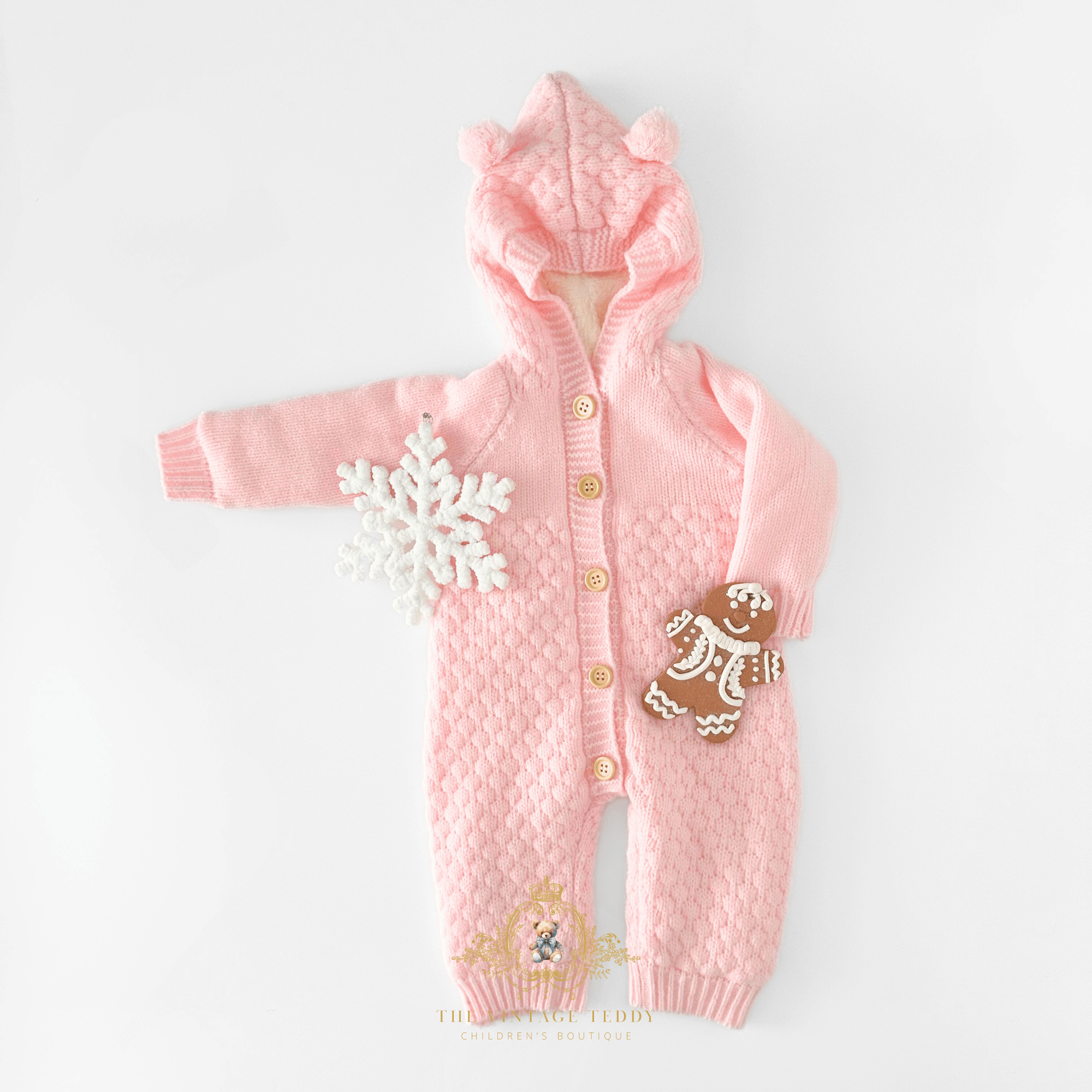 A pink knitted baby hooded romper with tan buttons, styled with a white plush snowflake on one sleeve and a gingerbread cookie ornament placed on the leg, laid flat on a white background.