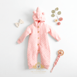 Pastel pink knitted baby one-piece sweatsuit with a hoodie, front button closure, and teddy ear details on the hood, laid flat on a white background with decorative props including a bow, flowers, and a crochet pacifier holder.