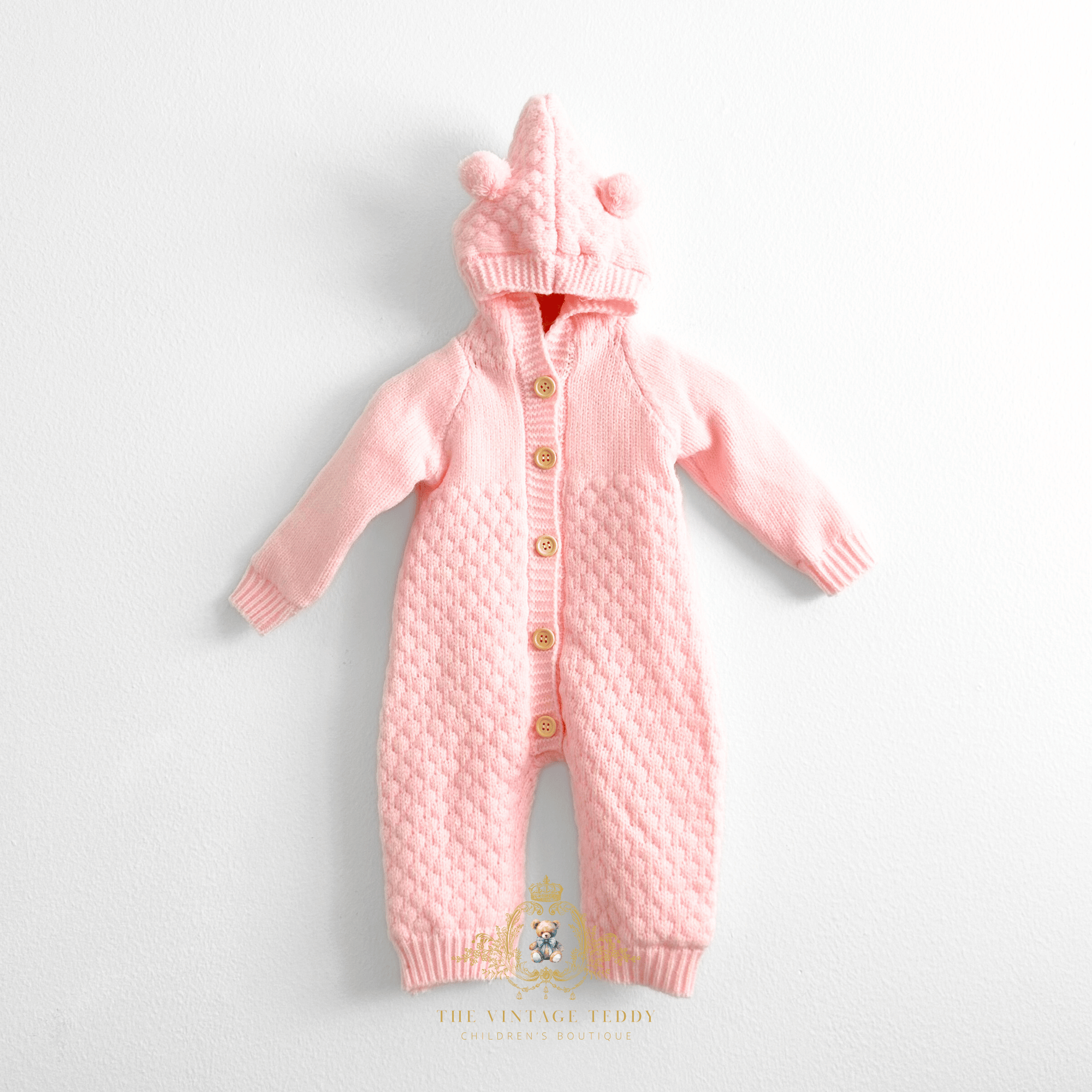 Pastel pink knitted baby jumpsuit with a hood featuring two bear ears and three wooden buttons, fleece lining laid flat on a white background. A watermark at the bottom reads The Vintage Teddy Children's Boutique.