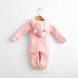 Pastel pink knit baby jumpsuit with a hoodie and cute teddy bear pom-pom details, hanging on a light natural wood hanger displaying the back of the outfit against a plain white wall.