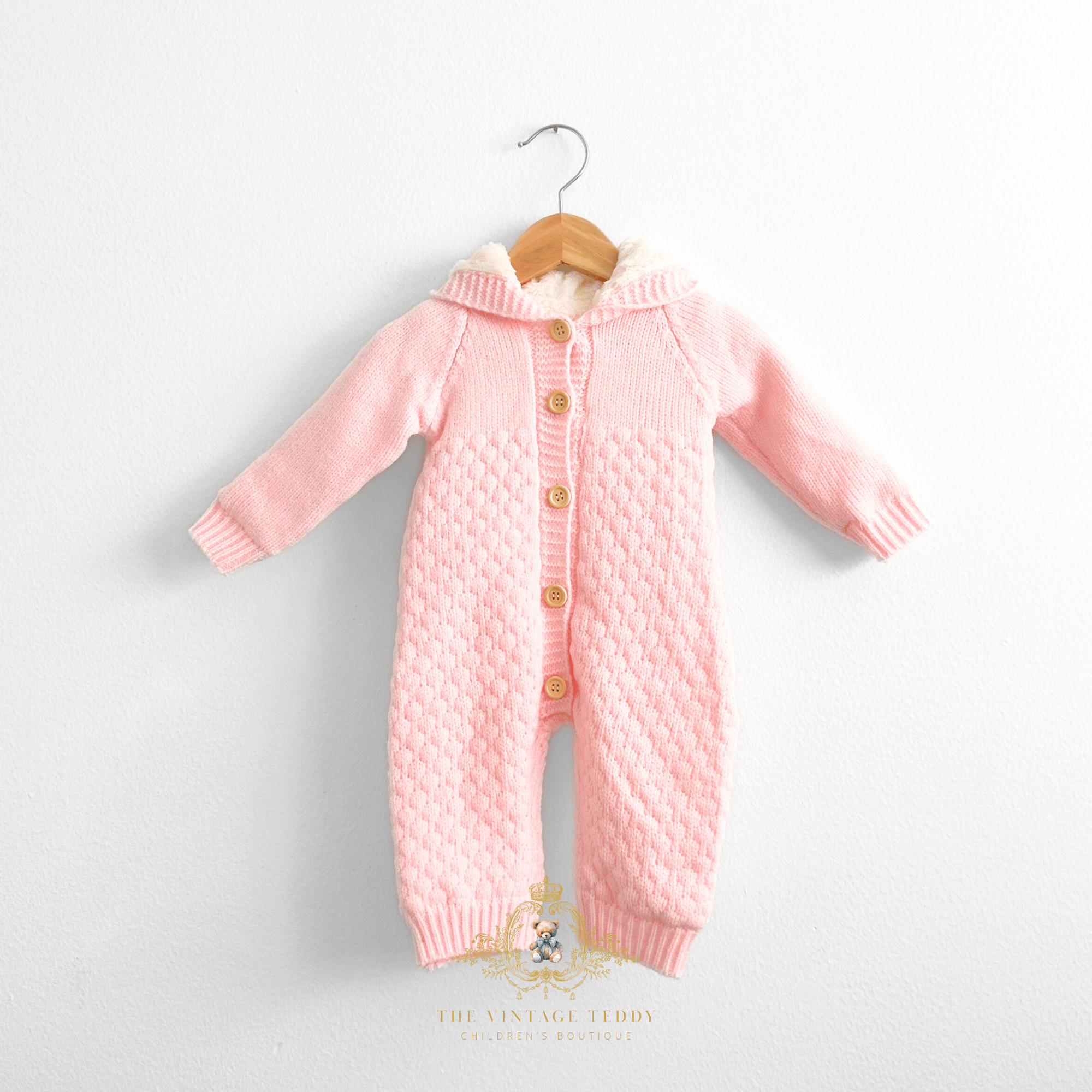 Baby pink knitted hooded jumpsuit for Baby & Toddler girls with delicate quilted pattern, tan buttons, and a cream-colored fleece plush lining inside the bodysuit and hood, hanging on a natural wooden hanger against a white wall. The Vintage Teddy watermark stamp at the bottom center.