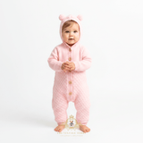 An adorable baby girl wearing a pink knitted hooded jumpsuit with bear ears on the hood and tan buttons down the front, standing against a plain white studio background. The Vintage Teddy logo mark at the bottom center.