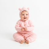 A smiling baby girl sitting wearing a pastel  pink knitted hooded baby one-piece with teddy bear pom pom ear details on the hood and front buttons, clean white studio background. A watermark at the bottom reads The Vintage Teddy Children's Boutique.