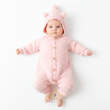 Baby girl lying on her back, top view,  wearing a pink knitted hooded jumpsuit with button-front closure and pom-pom bear ears on the hood, against a clean white background. 'The Vintage Teddy' children's boutique logo mark at the bottom center.