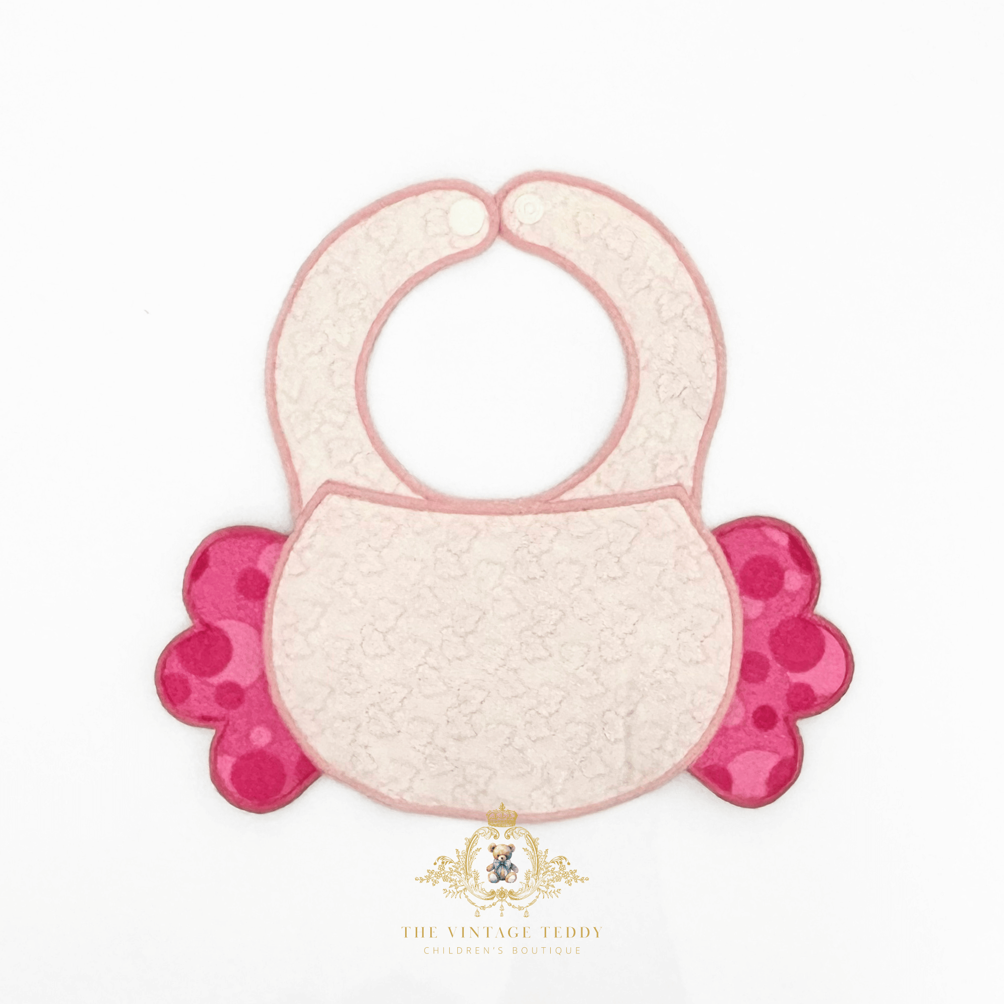 The back of an embroidered boutique baby bib with a pale pink textured center, a pink border, and bright pink scalloped side flaps, shown on a white background with a decorative The Vintage Teddy's brand watermark below.