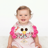A smiling baby girl wearing a pink dress and a decorative patchwork boutique baby bib, sitting against a clean white background. The bib features a n embroidered owl face with a yellow beak, pink polka-dot wings, floral fabric and a plaid neckband. The Vintage Teddy brand watermark is visible at the bottom.