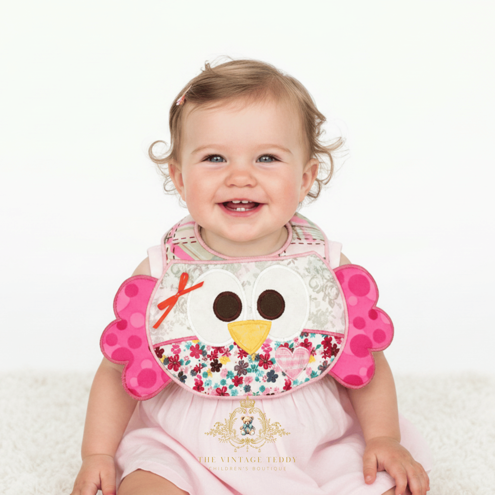 A smiling baby girl wearing a pink dress and a decorative patchwork boutique baby bib, sitting against a clean white background. The bib features a n embroidered owl face with a yellow beak, pink polka-dot wings, floral fabric and a plaid neckband. The Vintage Teddy brand watermark is visible at the bottom.