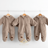 Three hooded fleece baby jumpsuits in a warm taupe-brown color, hanging on wooden hangers from a white rod against a white wall. Each kids jumpsuit has long sleeves and embroidered paw print details on the legs