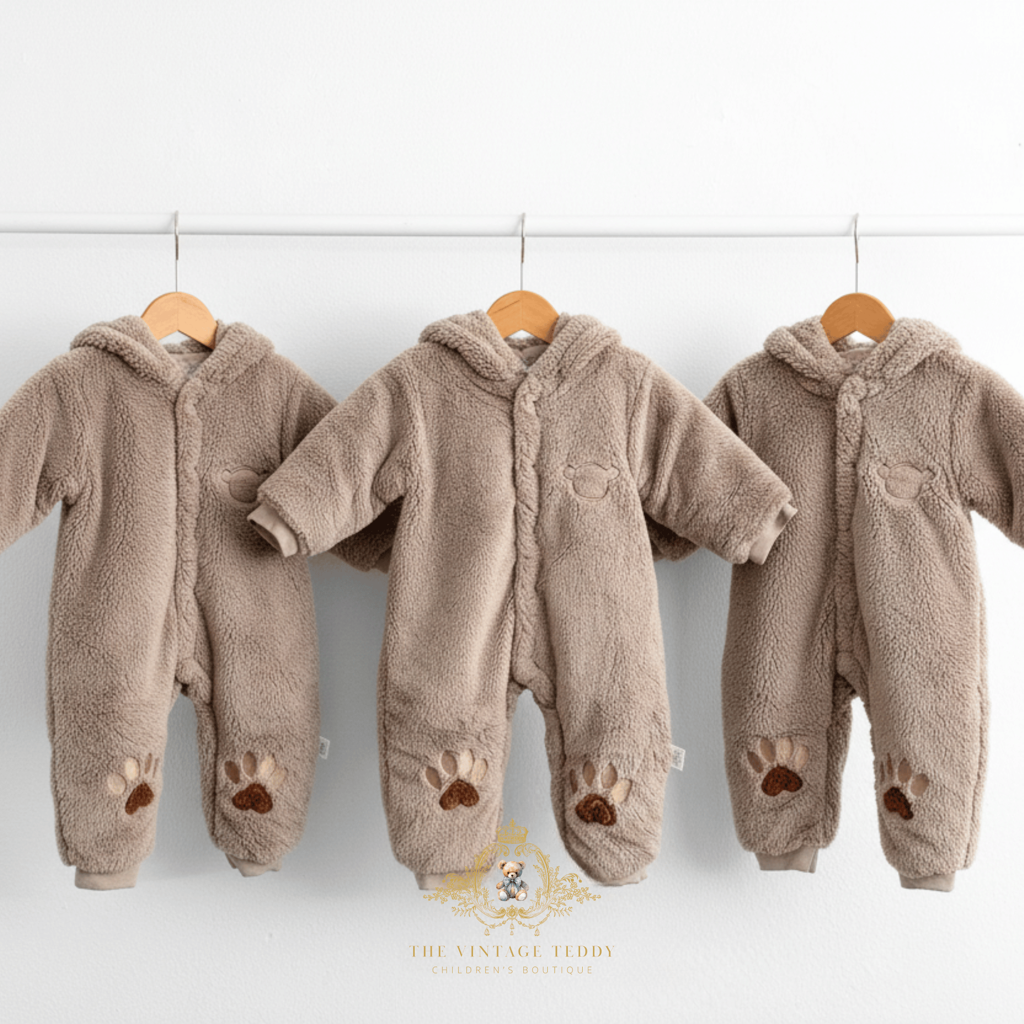Three hooded fleece baby jumpsuits in a warm taupe-brown color, hanging on wooden hangers from a white rod against a white wall. Each kids jumpsuit has long sleeves and embroidered paw print details on the legs