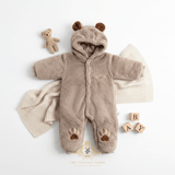 A baby fleece hooded one-piece outfit in warm taupe, styled flat-lay with a cream knit blanket, a small teddy bear, and wooden letter blocks on a white background. The outfit has brown bear ears on the hood, an embroidered teddy bear detail on the chest and paw prints on the legs.