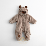 A baby's plush hooded one-piece romper in a warm taupe-brown color, designed to look like a teddy bear. It features brown faux-fur ears on the hood, a front snap button closure, and paw-print embroidered details on the the legs.