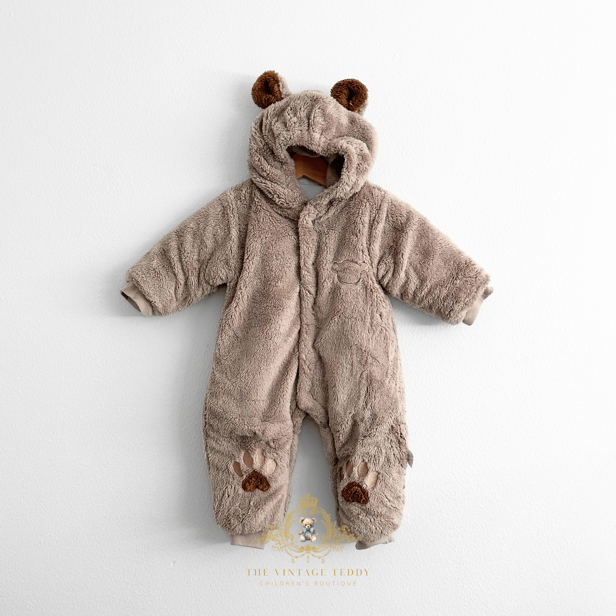A baby's plush hooded one-piece romper in a warm taupe-brown color, designed to look like a teddy bear. It features brown faux-fur ears on the hood, a front snap button closure, and paw-print embroidered details on the the legs.