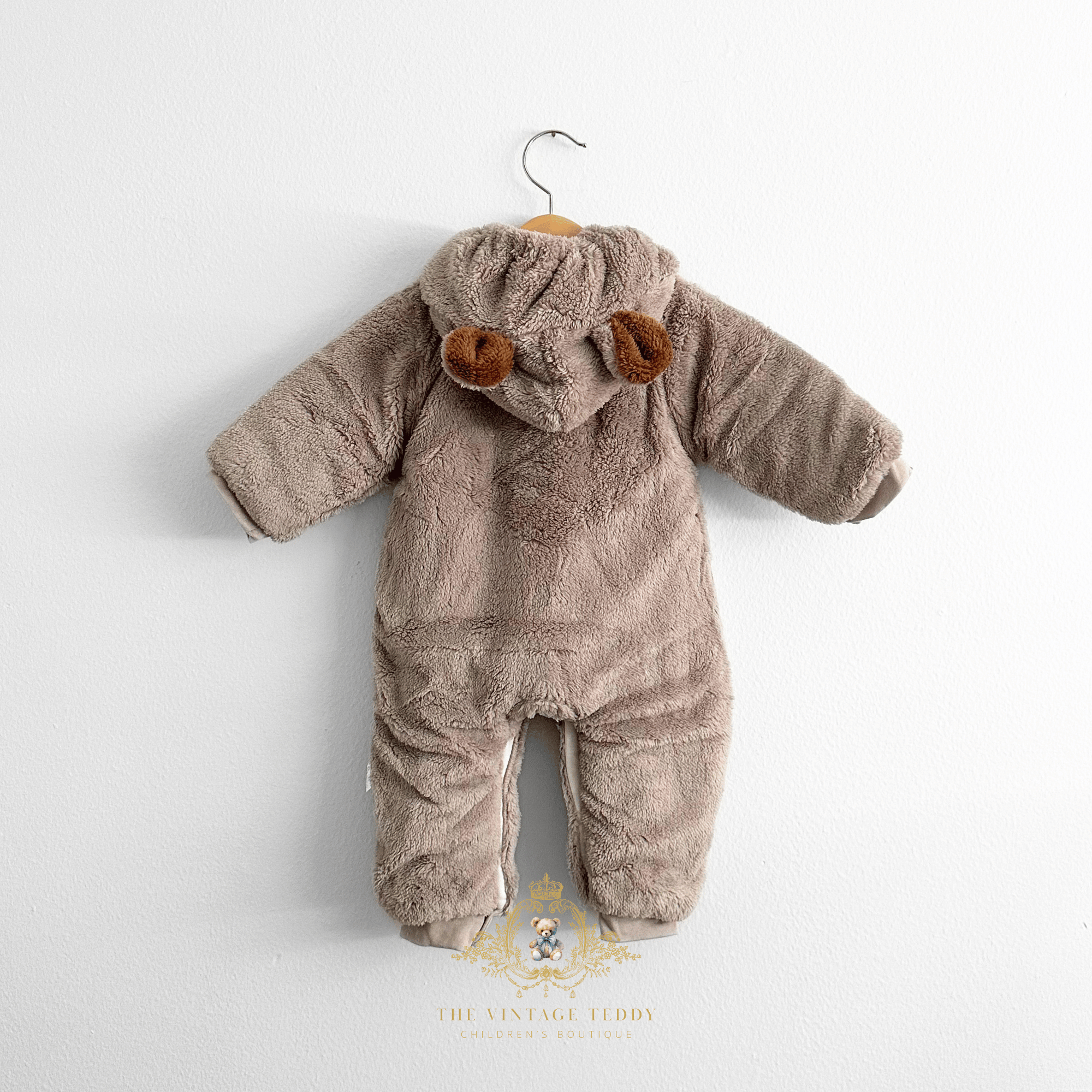 Baby & Toddlers hooded one-piece romper in a soft taupe-brown plush fleece fabric, hanging on a wooden hanger, against a plain white wall. The hood features two small brown faux-fur ear accents, and the romper has long sleeves and footless leg