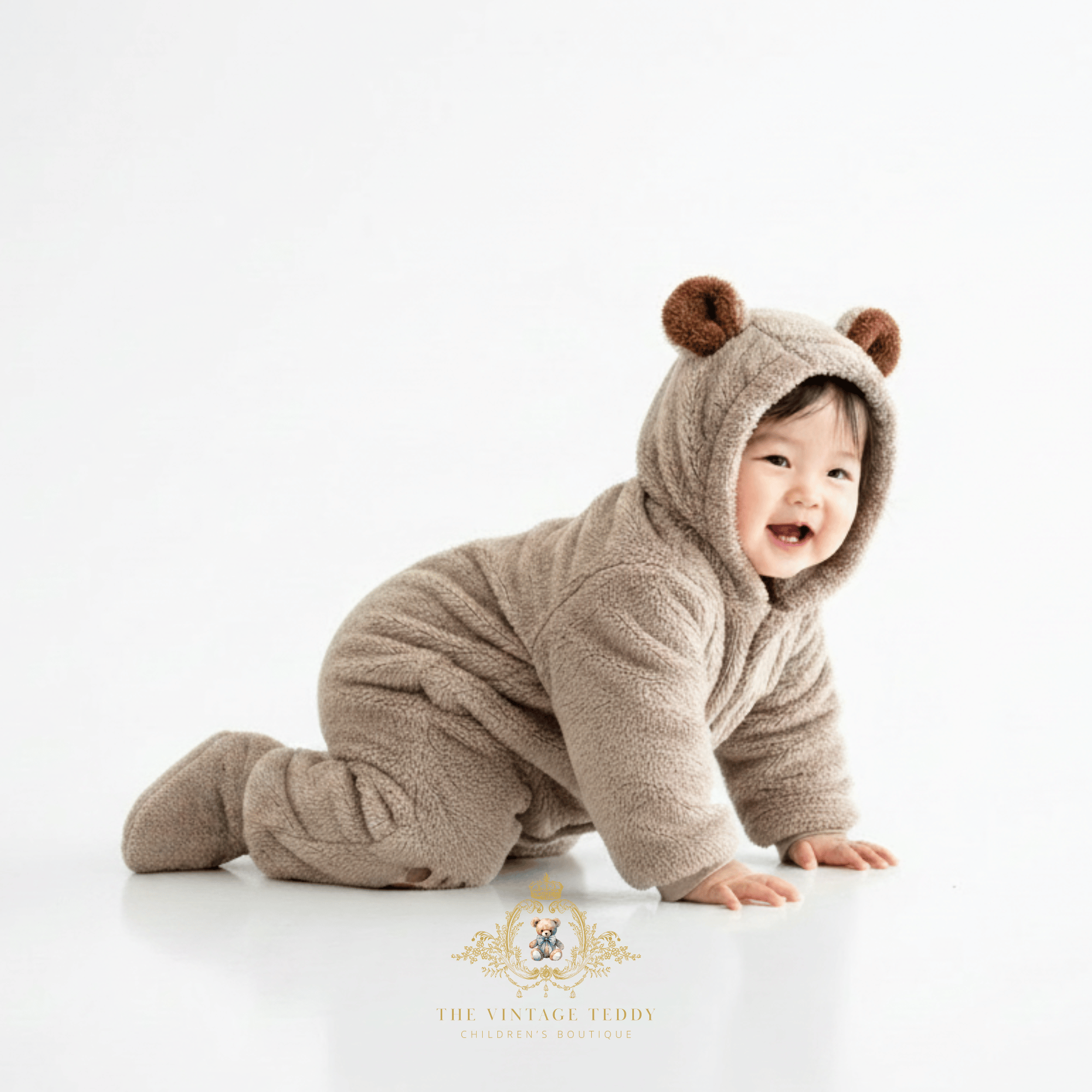 Baby boy crawling on a white studio floor wearing a plush hooded one-piece outfit with bear ears and footies. The garment is a warm taupe-brown color, and a watermark logo for 'The Vintage Teddy Children's Boutique' is visible at the bottom.