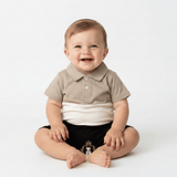 A smiling baby sitting on a white studio background, wearing a short-sleeve polo one-piece romper with a beige collar and upper chest, a white horizontal band, and black shorts.
