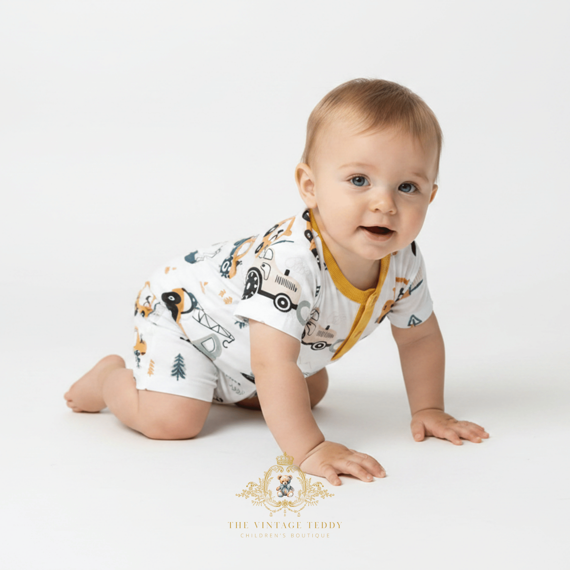 A baby crawling on a white studio floor wearing a white short-sleeve romper with a yellow collar and a playful construction themed print. The brand logo 'The Vintage Teddy Children's Boutique' is visible at the bottom.