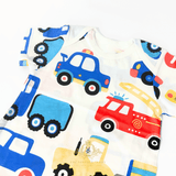 A close-up of a white short-sleeve baby romper featuring a colorful cartoon pattern of vehicles, including blue police cars, a red fire truck, and a yellow construction truck. The shirt is laid flat on a white background with a The Vintage Teddy's  watermark bottom.