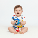 A baby sitting on a white floor against a white background, wearing a white short-sleeve romper with a colorful toy truck print and holding a blue toy cup. The brand name 'The Vintage Teddy' is visible at the bottom.