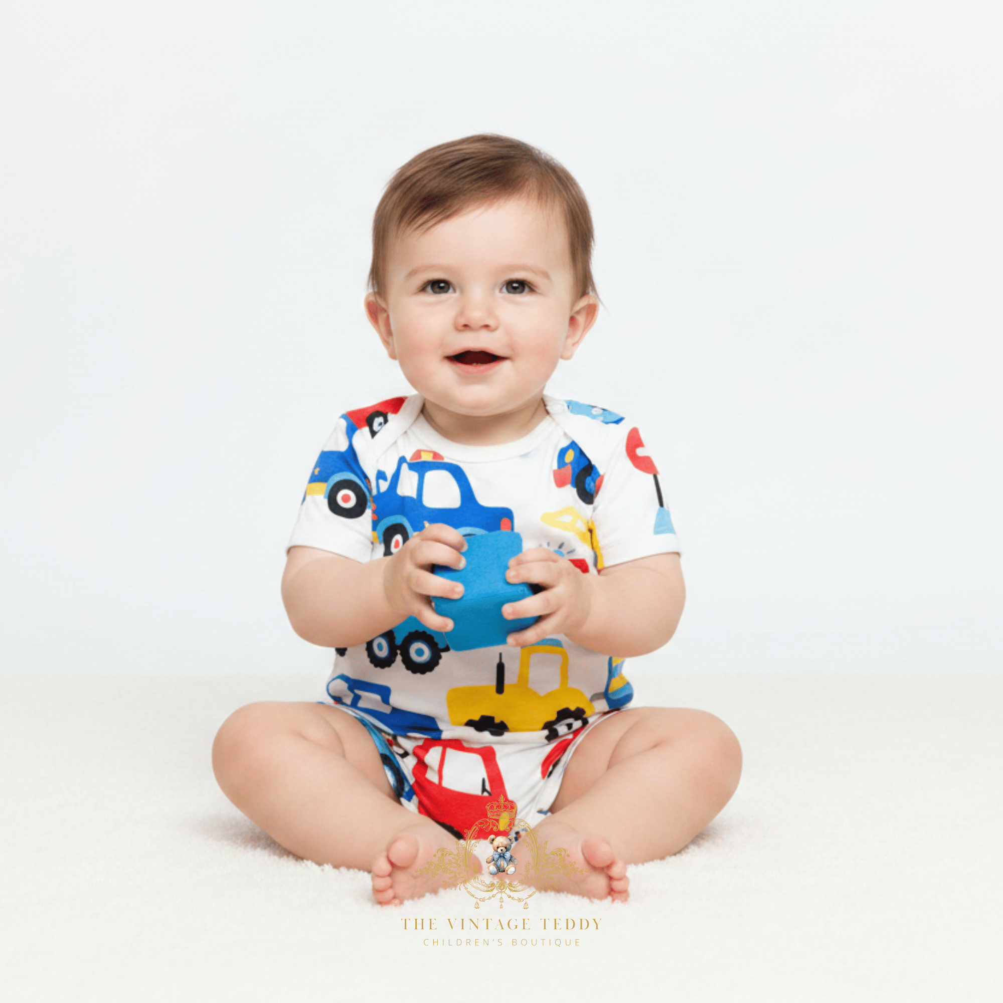 A baby sitting on a white floor against a white background, wearing a white short-sleeve romper with a colorful toy truck print and holding a blue toy cup. The brand name 'The Vintage Teddy' is visible at the bottom.
