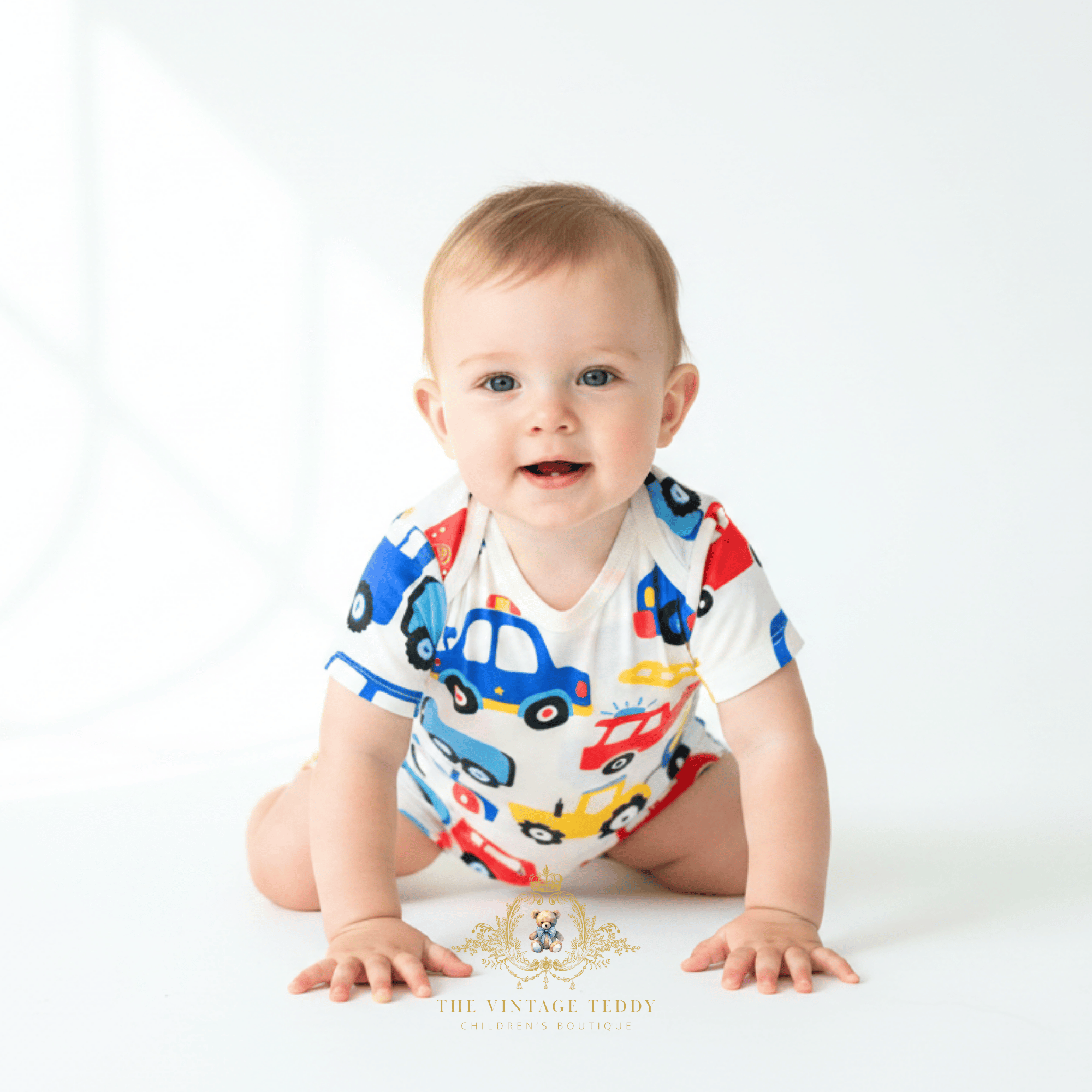 A baby wearing a white short-sleeve bodysuit with a colorful car print, crawling on a white studio background. The brand logo 'The Vintage Teddy Children's Boutique' is visible at the bottom.