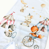 A close-up of a white baby garment printed with whimsical circus-themed watercolor animal illustrations, including a fox on a penny-farthing, an elephant on a scooter, a rabbit swinging from a moon, balloons, stars, and striped tents.