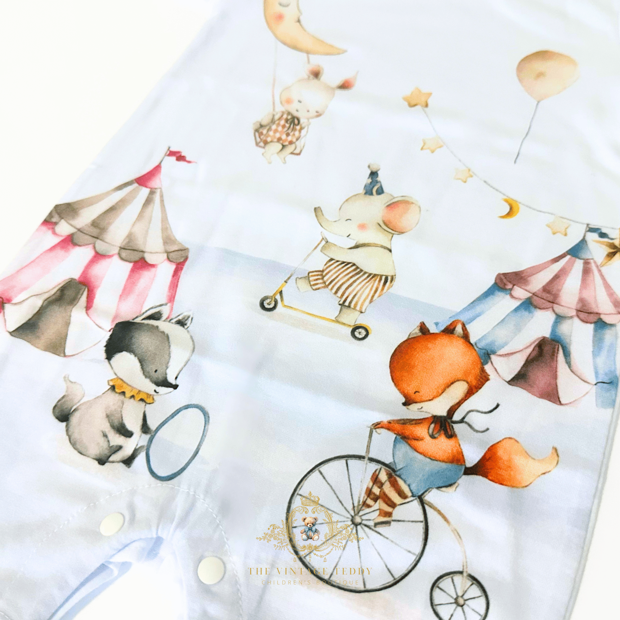 A close-up of a white baby garment printed with whimsical circus-themed watercolor animal illustrations, including a fox on a penny-farthing, an elephant on a scooter, a rabbit swinging from a moon, balloons, stars, and striped tents.