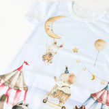 A close-up of a white short-sleeve baby one-piece romper featuring a whimsical watercolor carnival print of a bunny on a swing under a crescent moon, an elephant on a tricycle, stars, balloons, and circus tents.