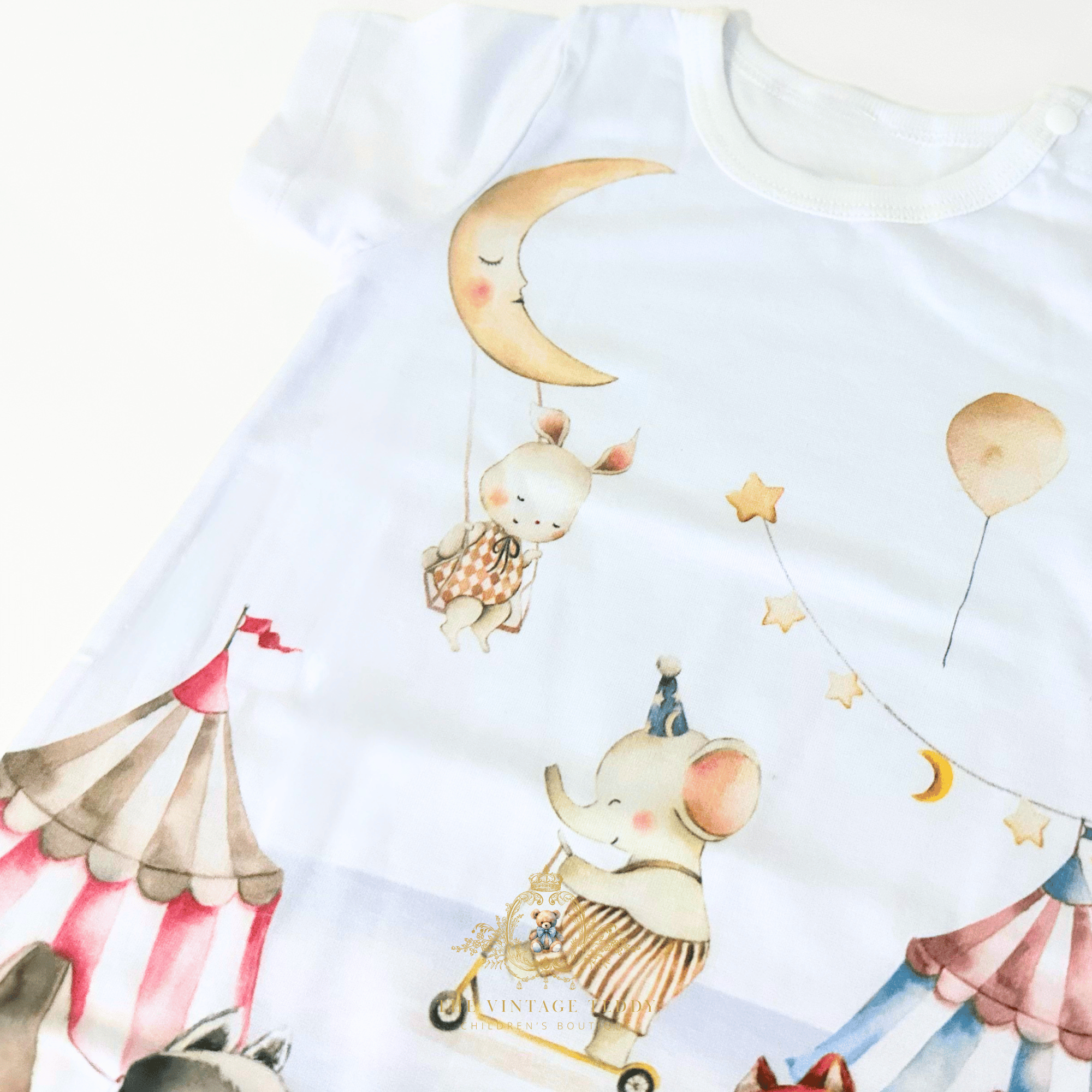 A close-up of a white short-sleeve baby one-piece romper featuring a whimsical watercolor carnival print of a bunny on a swing under a crescent moon, an elephant on a tricycle, stars, balloons, and circus tents.
