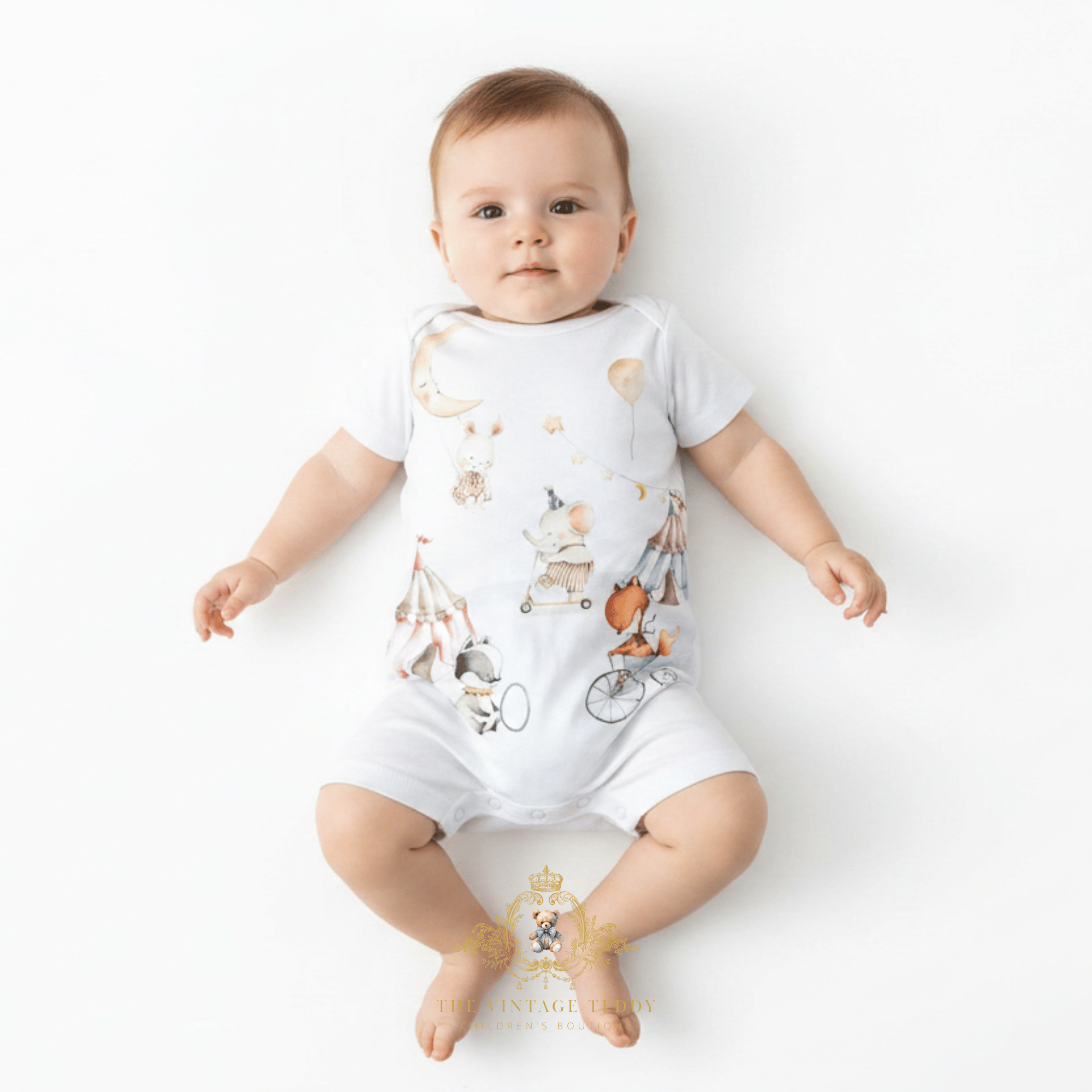 A baby lying on their back wearing a white short-sleeve bodysuit with a whimsical cartoon print of a moon, hot air balloon, bicycle, and tent. The photo is taken on a clean white background with The Vintage Teddy's watermark at the bottom.