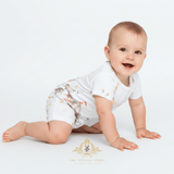 A smiling baby crawling on a white studio floor wearing a white short-sleeve bodysuit baby romper with a delicate multicolor carnival graphic print design. The brand logo 'The Vintage Teddy Children's Boutique' is visible at the bottom.