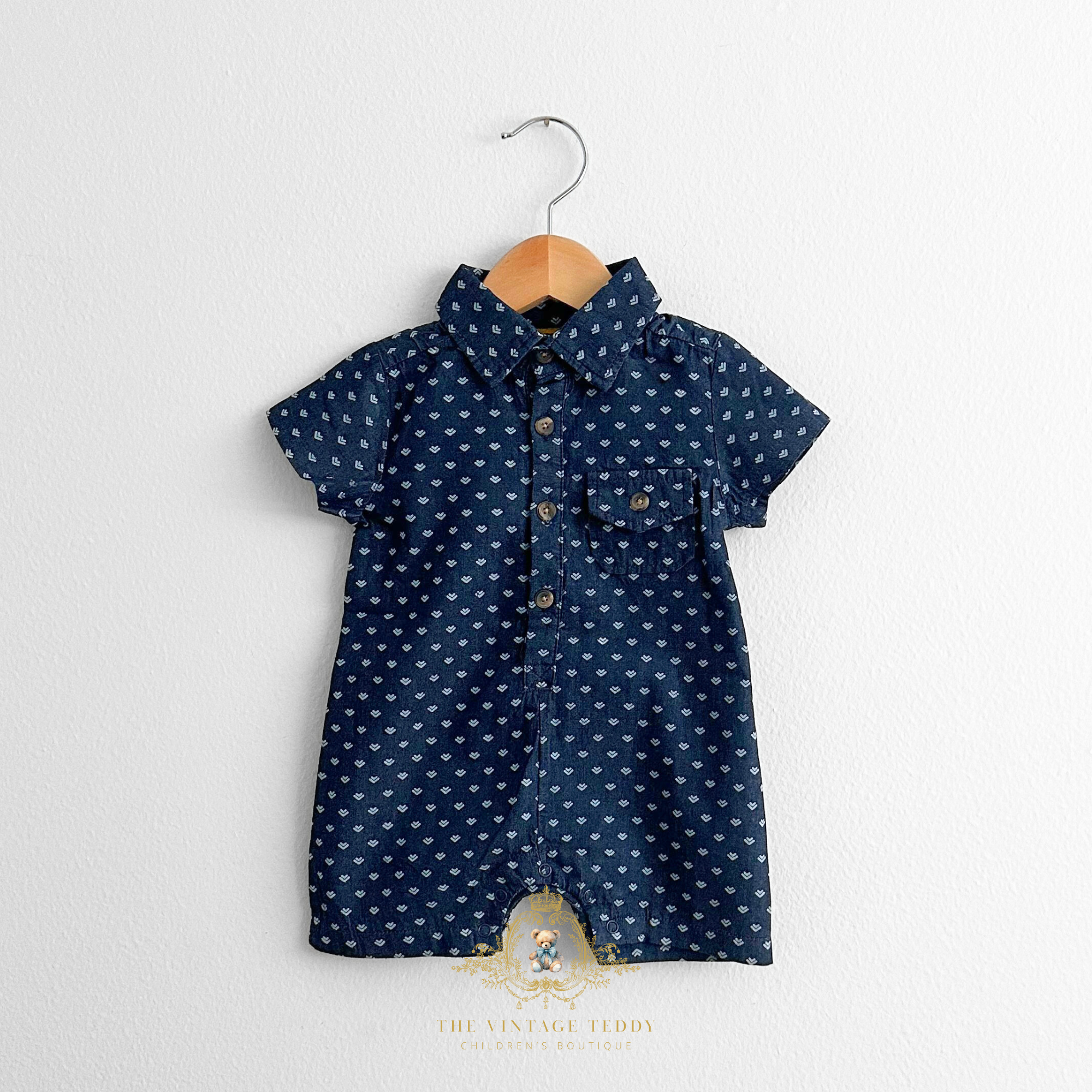 Adorable navy blue and arrows print boys one-piece romper on a wooden hanger against a white wall background. Baby & Toddler Clothes from The Vintage Teddy children's clothing store.
