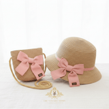 A kids straw sun hat and matching woven crossbody bag in beige, both decorated with large dusty pink bows and pink bands, displayed on a white surface against a white background. A logo for 'The Vintage Teddy' is visible at the bottom.