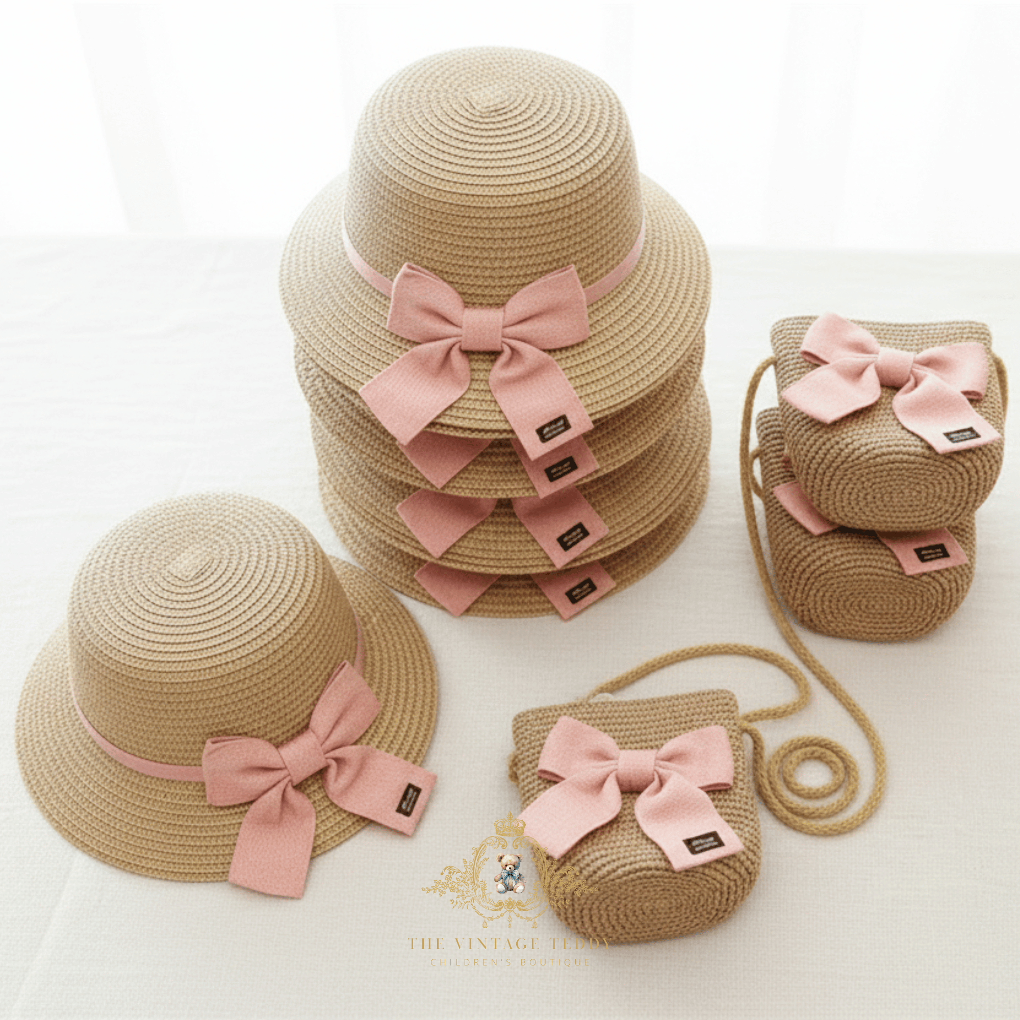 A coordinated set of children's woven straw hat and matching crossbody bags in a natural tone, each decorated with a soft pink bows. Stacks of sun hats, and shoulder /crossbody bags, arranged on a light fabric surface.