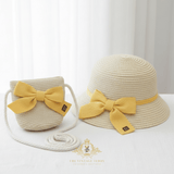 A straw bucket hat and matching woven shoulder bag set for children in a light beige color, both featuring a prominent yellow fabric bow with a small black brand tag. The bag has a thick white rope strap coiled in front.